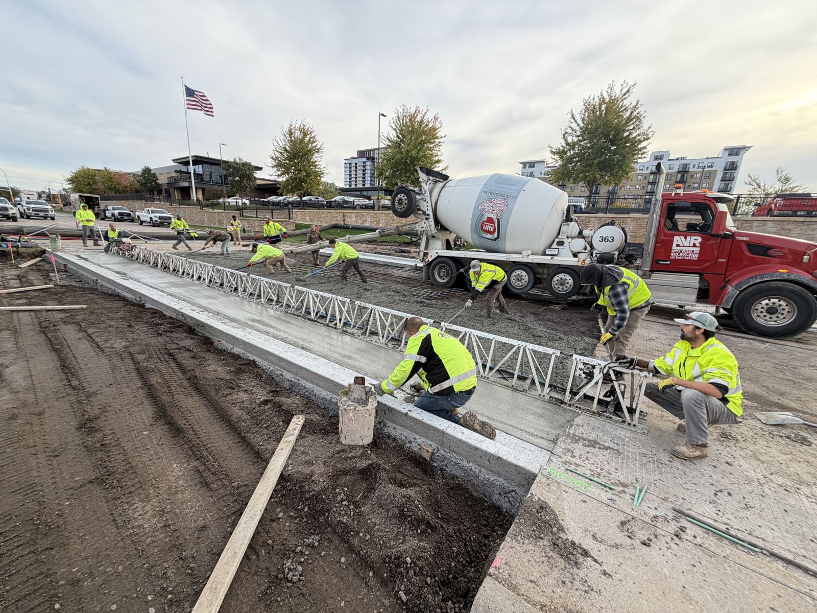 Concrete crew placing and screeding a pavement pour beside a new curb line, with a ready-mix truck