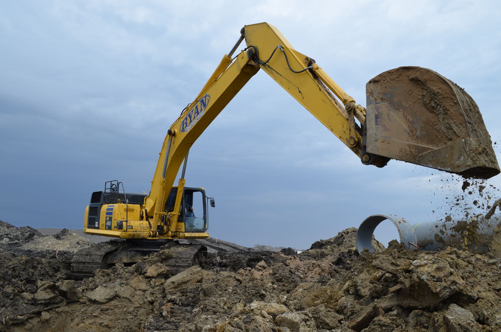 A Ryan Contracting excavator dumping a bucket of earth beside a trench, with large storm pipe behind it