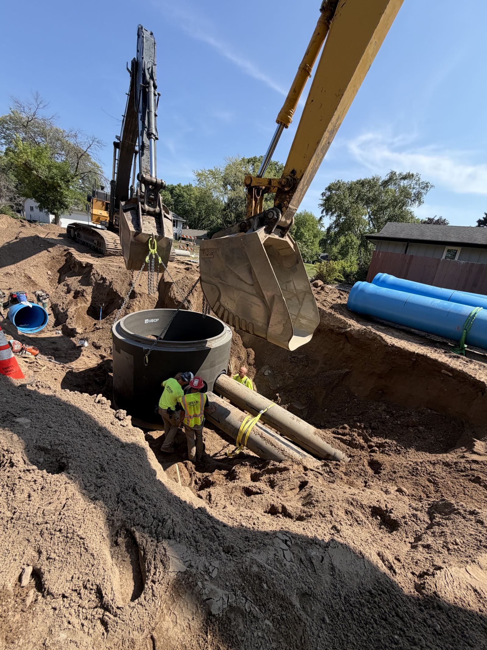 Pipe crew setting a manhole structure with new watermain staged beside the trench