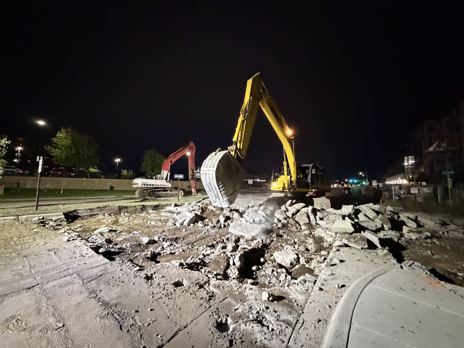 Excavator breaking out old pavement at night under work lights