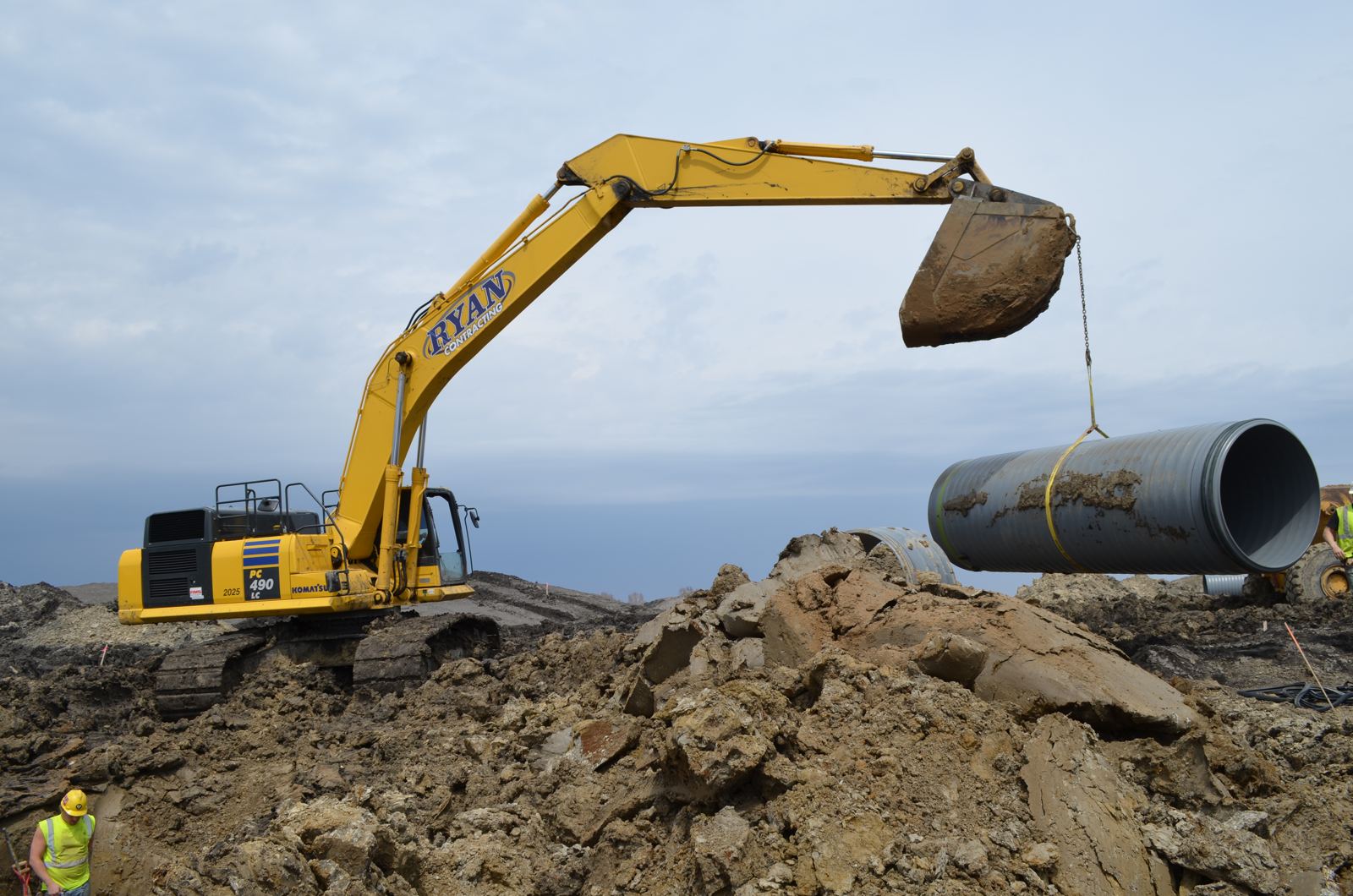 Excavator swinging a length of large-diameter pipe on a sling