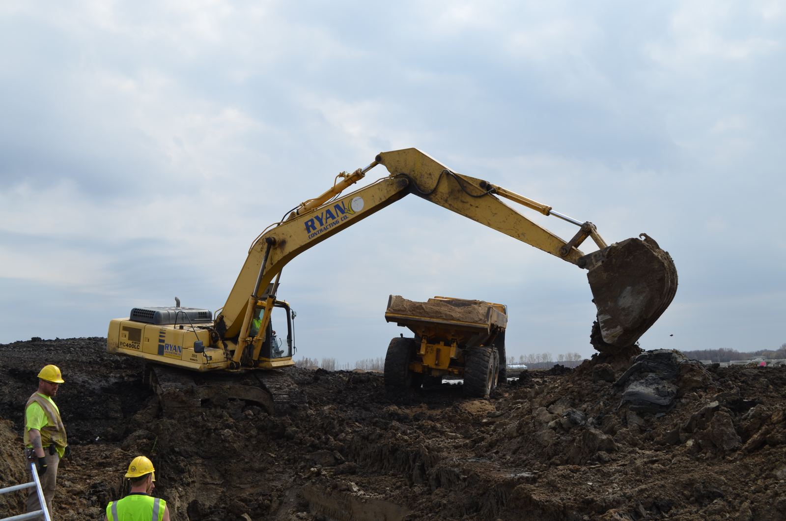 Excavator loading an off-road haul truck while crew works in the trench below